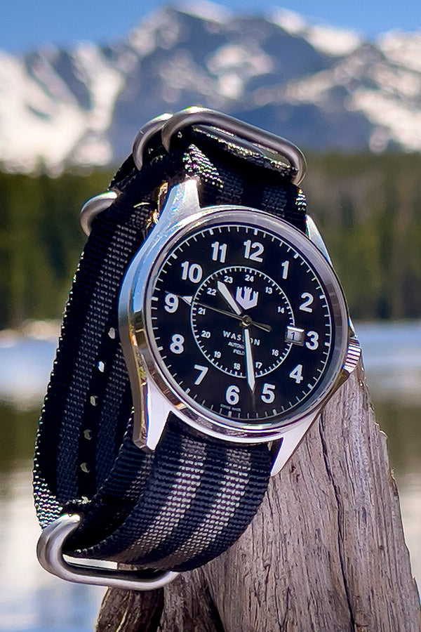 A Wasson Automatic Field Watch with a black and grey striped nylon strap sits propped on an old piece of driftwood. You can see a lake and snow-capped mountains in the background.