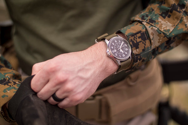 Close up image of a man's midsection. He is wearing fatigues and a Swiss made watch with an olive green nylon strap that meets all combat standards. He is holding a military backpack.