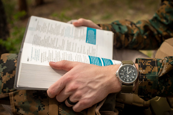 Closeup of a man wearing fatigues and a Wasson Automatic Field watch with an olive green nylon strap. He is holding an open Bible in his lap.