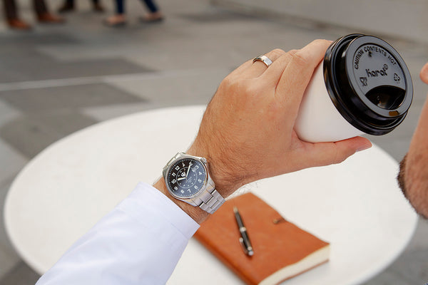 A closeup of a doctors left arm as he raises a paper coffee cup to his lips. He wears a stainless steel, Swiss-made Wasson Automatic Field Watch. He sits outside at a small table with a journal and pen on the table. 