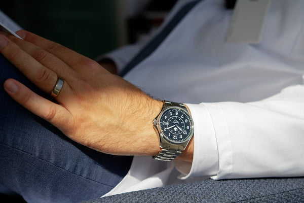 A closeup of a doctor sitting in a blue armchair reading a book. You can see only his torso clad in a white lab coat. He wears a stainless steel Wasson Automatic Field Watch on his left wrist.