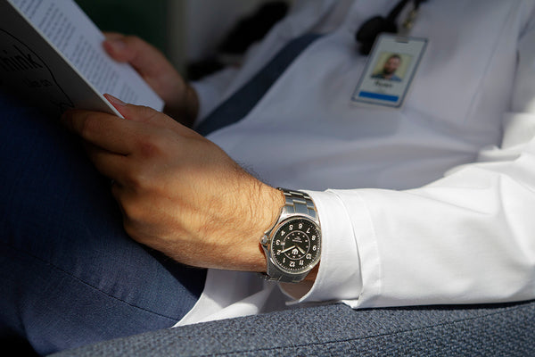 A closeup of a doctor sitting in a blue armchair reading a book. You can see only his torso clad in a white lab coat. He wears a stainless steel Wasson Automatic Field Watch on his left wrist.