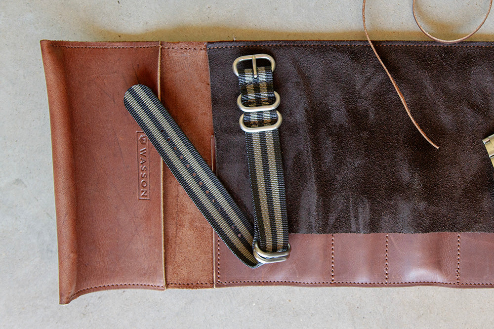 A black and gray zulu strap sits on top of a Wasson Watch leather tool roll.