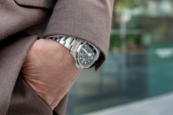 Closeup of a man in a brown suit with his hand in his pocket. You can see a stainless steel automatic watch beneath the cuff of his suit jacket.