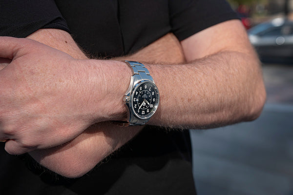 A very closeup image of a man's arms folded across his chest. He is wearing a black shirt and a Swiss-made Wasson Automatic Field Watch.