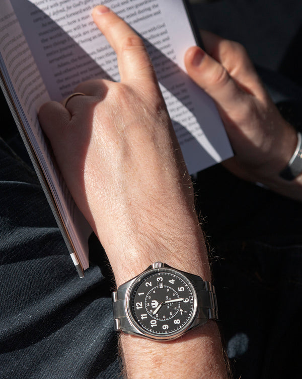Closeup image of a man's hand holding a book while wearing a Swiss-made Wasson Automatic Field Watch with a stainless steel case and bracelet.