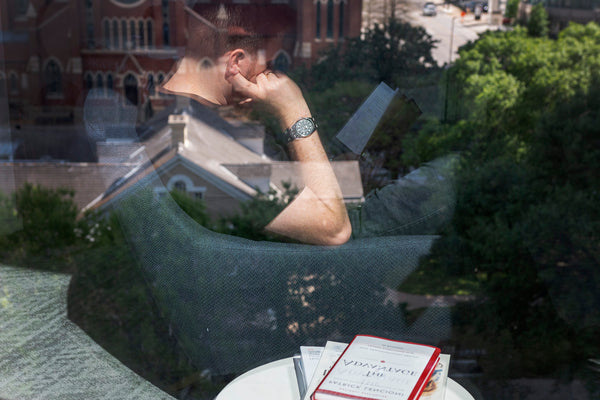 An abstract image of a man sitting in a lounge chair reading a book. It is shot through a window, so the glass reflects a city scape in the background. The man is reading a book while wearing a Wasson Automatic Field Watch.