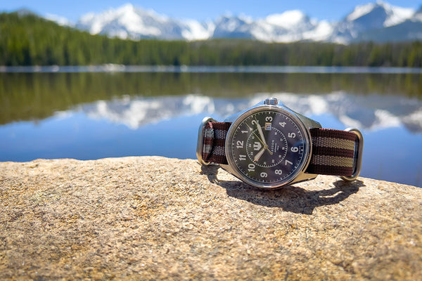 A Wasson Automatic Field watch with a maroon and gray striped nato strap sits atop a rock. There is a lake and mountains in the background.