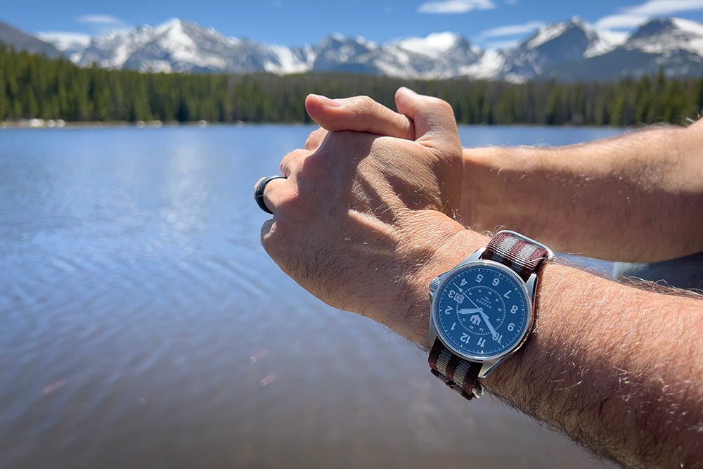 Closeup of a man's hands clasped together. He is wearing a Swiss made automatic field watch with a gray and maroon striped nato strap. In the background is a lake and snow-capped mountains.
