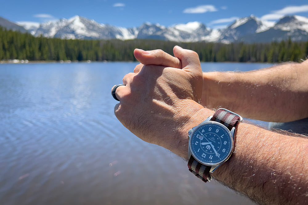 Closeup of a man leaning on a bridge wearing a Wasson Automatic Field Watch with a maroon and gray zulu strap. We see the lake and mountains in the background.
