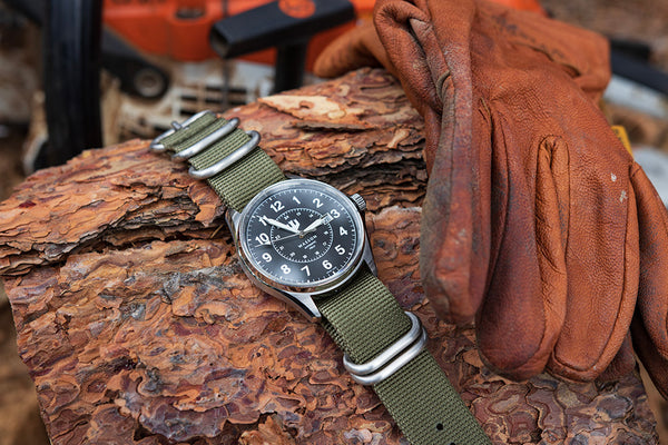 Swiss Made Automatic Field Watch with green nato strap sits on a log with leather gloves beside it and a chainsaw in the background.