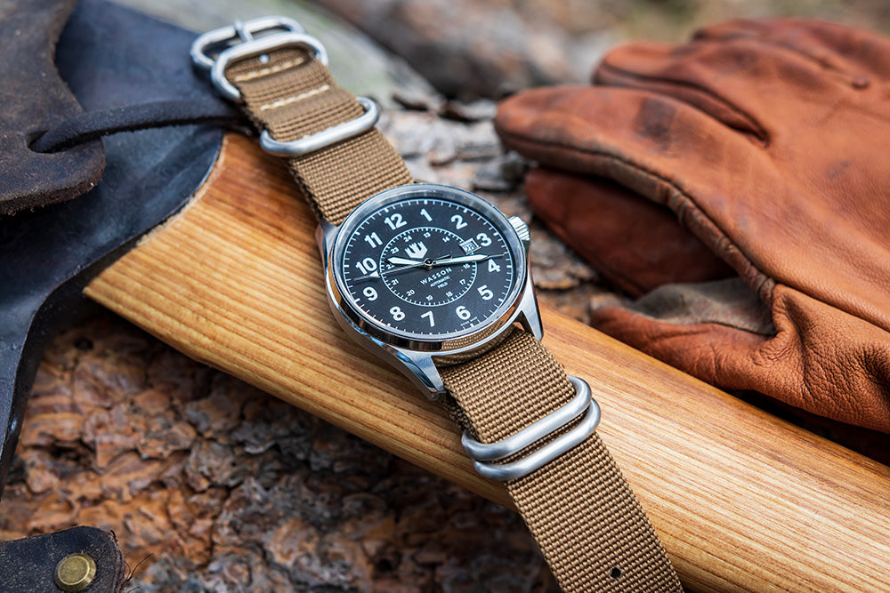 A Swiss made Field Watch with khaki nato strap is laid over a wooden axe handle. There are leather work gloves in the background.
