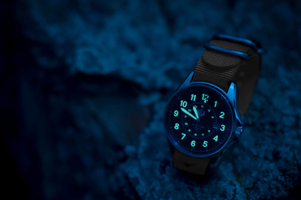A night shot of a Wasson Automatic Field watch with khaki nato strap sitting on a rock. The numbers and hands of the watch are glowing.