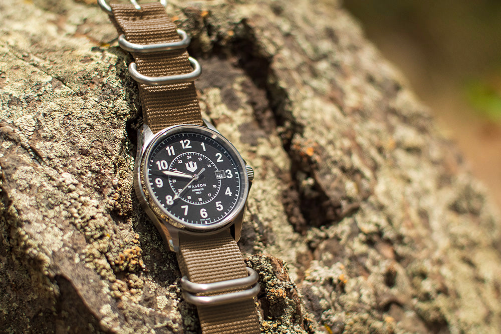A Wasson automatic field watch with a khaki zulu strap is laid over a textured rock.