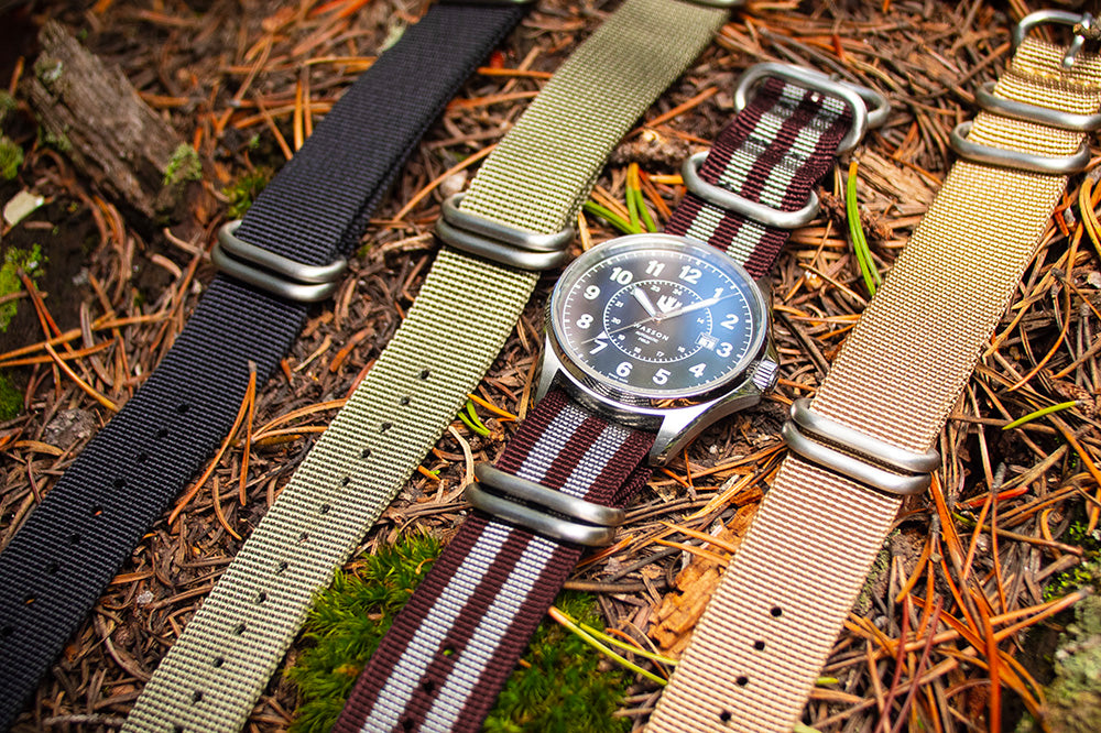 A Wasson automatic field watch with maroon and gray nato strap lays atop a bed of pine needles. There are three other nato straps beside it.