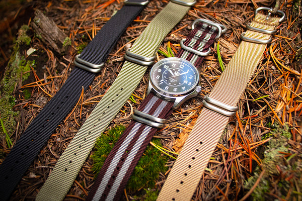 A Wasson Watch with a gray and maroon striped zulu strap lays on a bed of pine needles. There are three other zulu straps beside it.