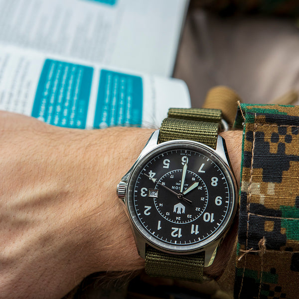 Extreme closeup of a man's wrist. He is wearing fatigues and a Wasson Watch Co. Automatic Field Watch with a nylon strap. He has an open book in his lap.
