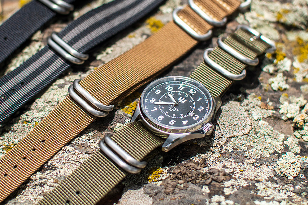 An Automatic Field Watch with an olive green zulu strap lays atop a textured rock. There are three other zulu straps in the background.