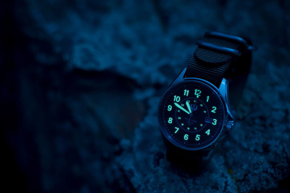A night shot of a Wasson Automatic Field Watch with a black nato strap sitting on a rock. The numbers and the hands of the watch are glowing.