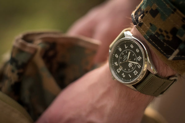 A military man in a camouflage jacket reaches into his backpack. He is wearing a Wasson Automatic Field watch with a green nato strap.