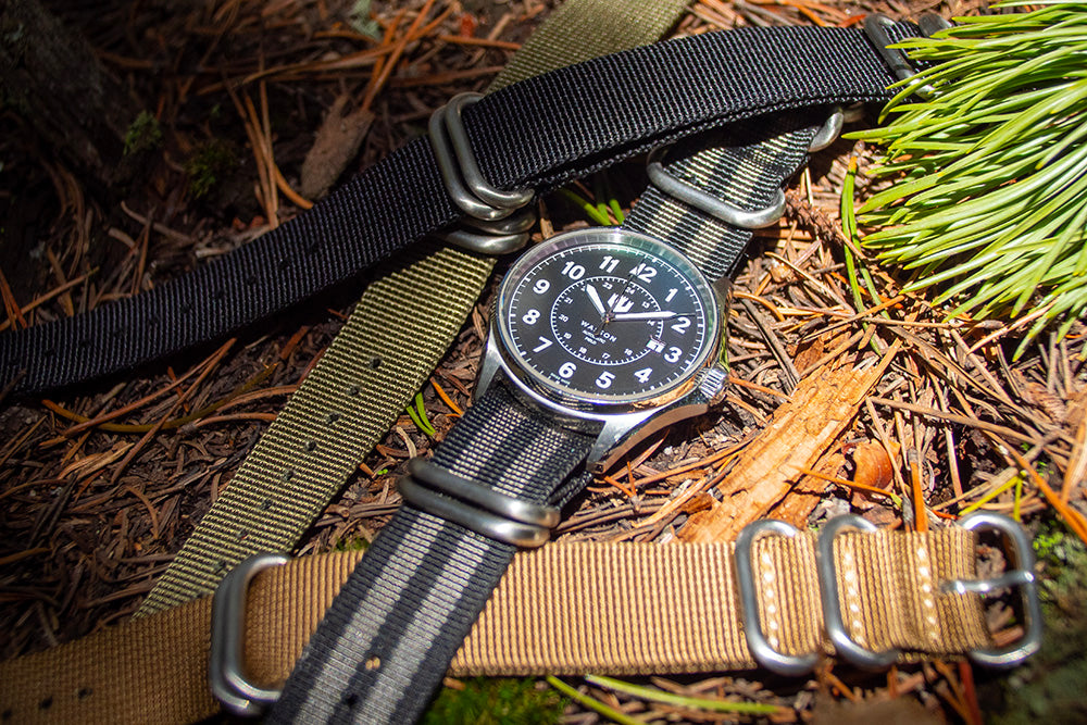 A Wasson Watch with black and gray zulu strap sits atop a bed of pine needles. There are three other zulu straps surrounding it.
