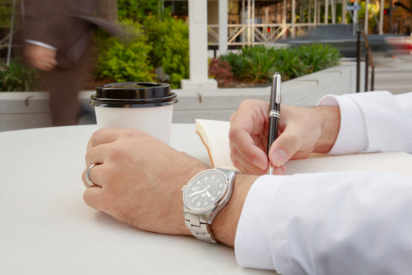 A closeup of a doctor's hands as he sits at a small table outdoors. He is holding a coffee in his left hand while wearing a Swiss made, stainless steel watch. He is writing in a journal with his right hand, and people are walking in the background.