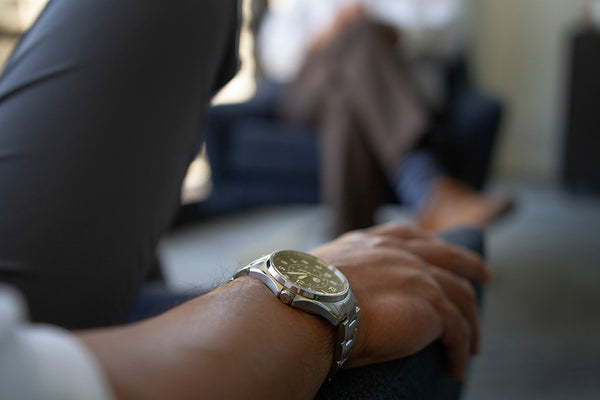 A closeup of a man's hand on the arm of an armchair while wearing a stainless steel automatic watch. You can see another man sitting in an armchair in the background.