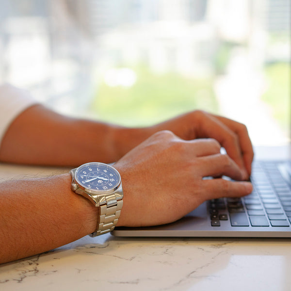 Closeup of a man's hands on the keyboard of a laptop. He is wearing a stainless steel, Swiss-made automatic field watch on his right wrist.
