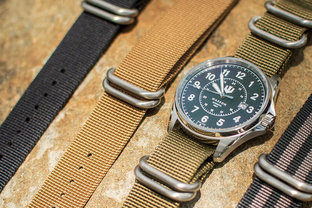 A Wasson Automatic Field Watch with a green zulu strap lays on a rock. There are three other straps laying on the rock beside it.