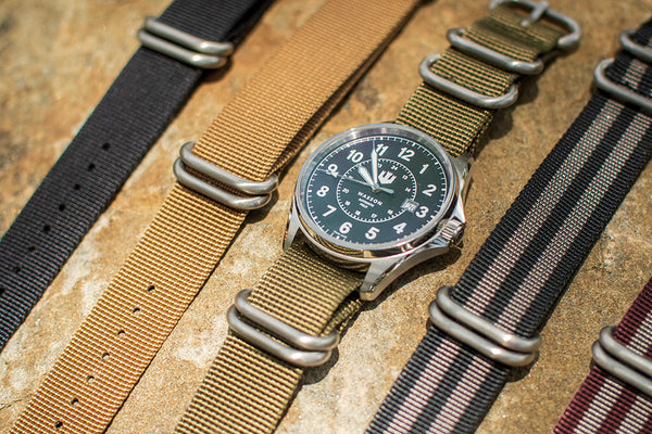 A Wasson Automatic Field Watch is shown with an olive green zulu strap laid out on a rock. There are four other zulu straps in various colors laying beside it.
