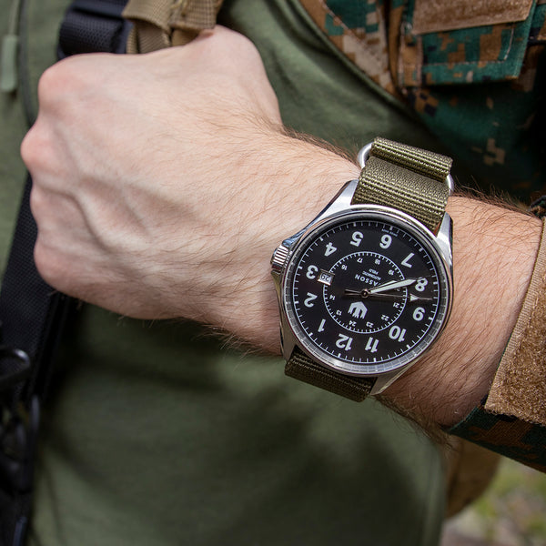 Close up of a man wearing fatigues and a Swiss made military watch with an olive green nylon strap wearing a backpack and holding its strap.