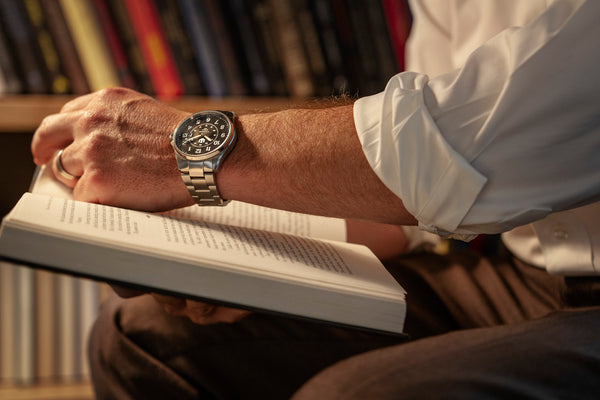 A closeup of a man holding a book in a library. He is turning the page with his left arm while wearing a Wasson Automatic Field Watch.