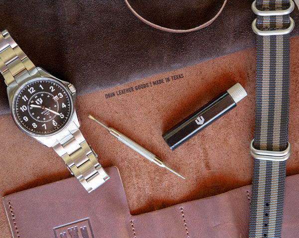 A closeup of a military field watch with a stainless steel bracelet lays on top of an open brown leather tool roll. A nylon strap and watch care tools sit beside the watch.