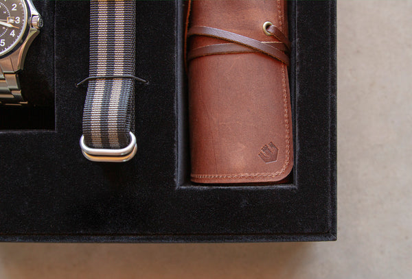 Closeup of a leather tool roll rolled up and nestled in a black gift box. The tool roll has the Wasson logo stamped on it. Beside that is a black and gray striped nylon bracelet and stainless steel Wasson Watch.