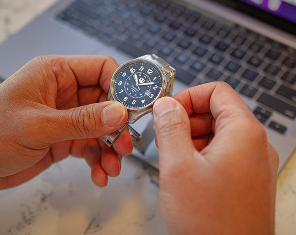 Closeup of a man holding a stainless steel watch in his hands. He is turning the crown to change the date or time. A laptop keyboard is seen in the background.