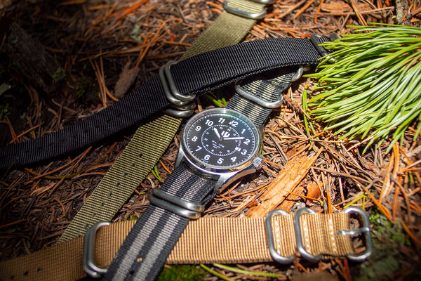 A Wasson Automatic Field Watch with a black and gray striped nylon strap lays on the ground amongst pine needles. There are three other nylon straps in khaki, black, and olive green laying with the watch.