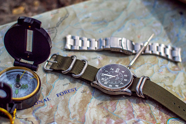 A Wasson Automatic Field Watch with an olive green nylon strap lays on a map of a national forest. Beside it is an open compass and a stainless steel watch band with a tool to change bands.