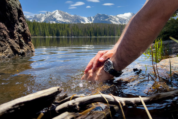 Closeup of a man washing his hands in a lake with pine trees and snowcapped mountains in the background. The man wears a water resistant Swiss made automatic field watch with a black nylon strap.
