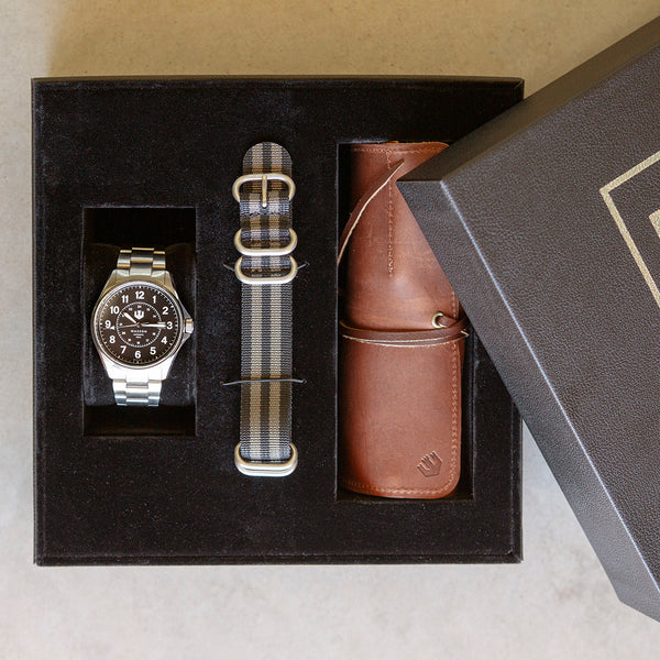 A Wasson Automatic Field watch is nestled in a black gift box with an extra nylon strap beside it and a leather tool roll rolled up inside the box, as well.