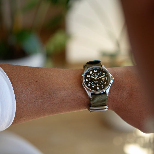 A closeup of a man with a raised wrist checking the time on his Swiss-made, stainless steel automatic watch with an olive green zulu nylon strap. The image is shot over his shoulder.