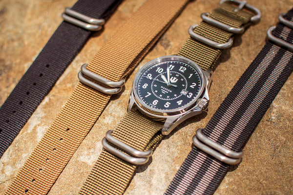 A Swiss-made automatic field watch with an olive green nylon watch band is laid on a tan colored rock. There is a black nylon strap, a khaki nylon strap, and a black and grey striped nylon strap laid out beside the watch.