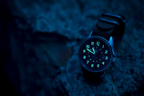 A Wasson Automatic Field watch sits propped on a rock late at night. The numerals and hands of the watch are glowing in the darkness.